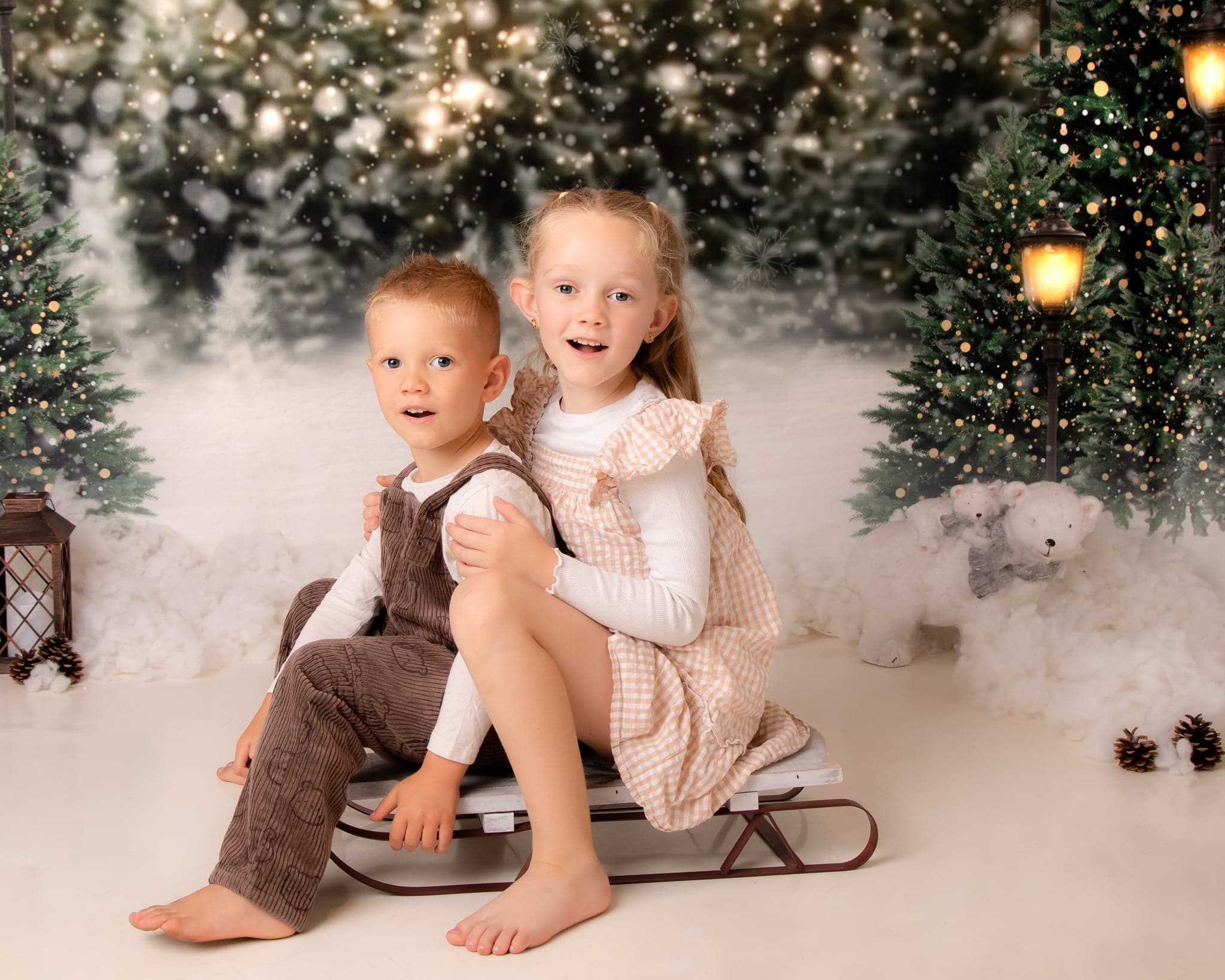 Two children sitting together among snowy Christmas trees in the studio.